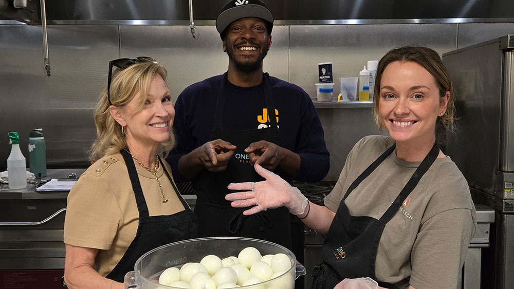 Three volunteers wearing aprons and gloves stand in a commercial kitchen, smiling while preparing food, with a large container of peeled hard-boiled eggs in the foreground.