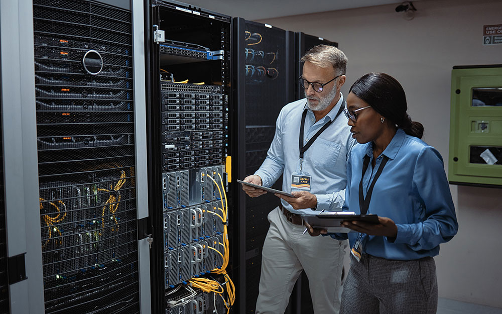 Two professionals wearing ID badges stand in a data center, reviewing information on tablets while inspecting server racks and network equipmen