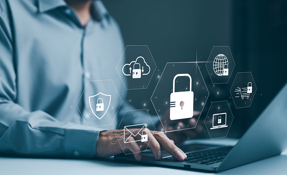 Person working on a laptop at a desk, with transparent digital security icons such as a padlock, shield, cloud, and email overlayed to represent cybersecurity and data protection.