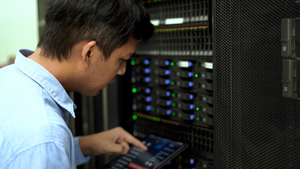 “IT technician inspecting server racks and monitoring system performance using a tablet.