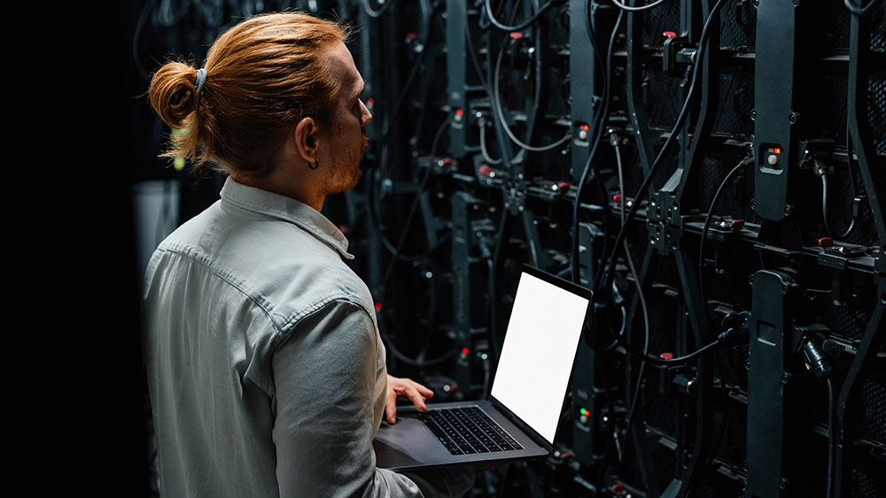 “IT technician inspecting server equipment while working on a laptop in a data center.