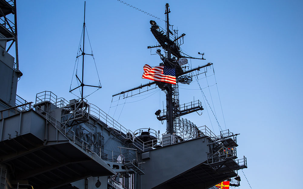 The upper deck of a U.S. naval ship is shown with radar and communication equipment, with an American flag flying against a clear blue sky.