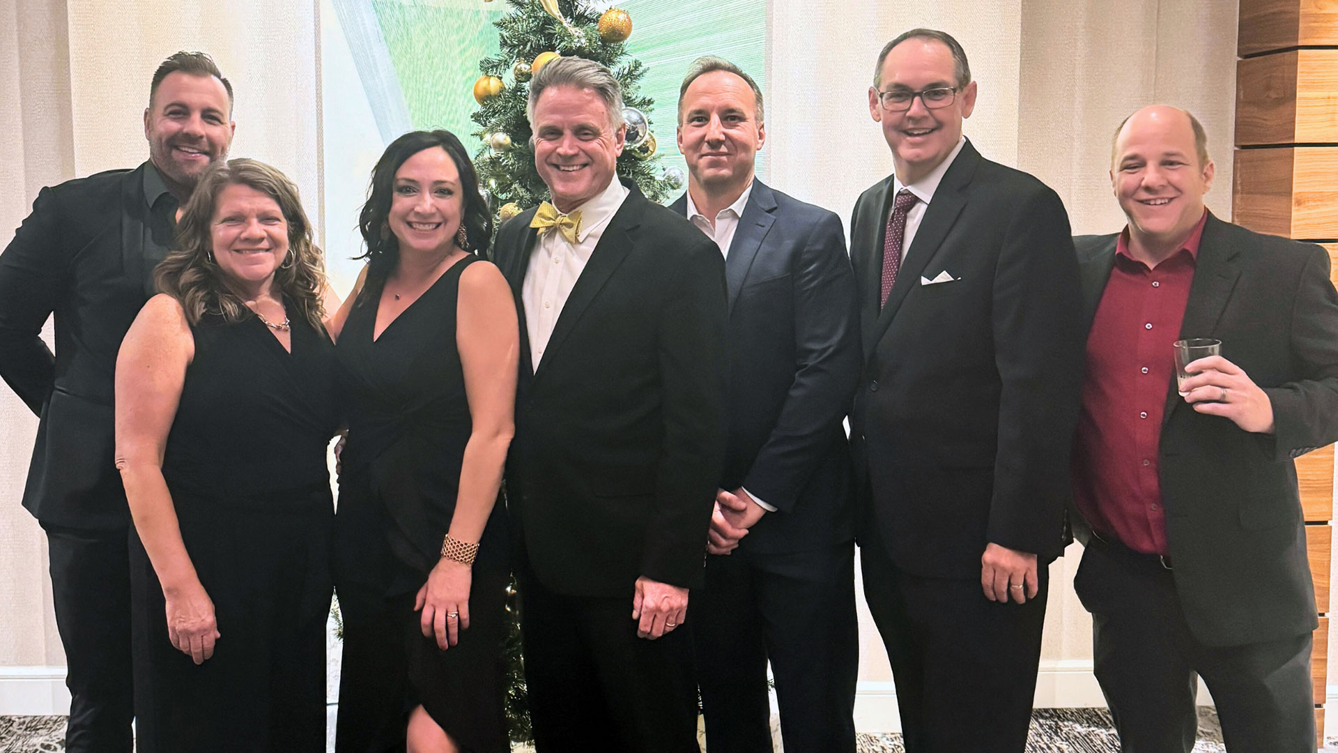 Group of eight professionally dressed adults stand together indoors in front of a decorated Christmas tree, smiling at the camera during a formal or corporate holiday event.