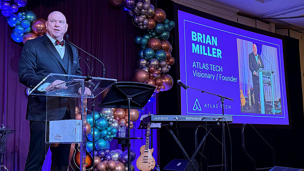 Man in a tuxedo speaks at a clear podium on a stage decorated with balloon arrangements, while a large screen beside him displays the text “Brian Miller, Atlas Tech, Visionary / Founder” along with his image during a formal event or awards ceremony.