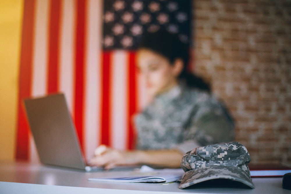 Student in uniform studying on a laptop, with an American flag displayed in the background.